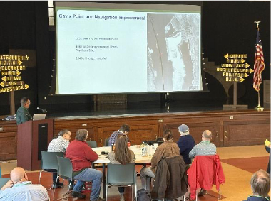 Person presenting in front of an audience looking at a large projection screen.